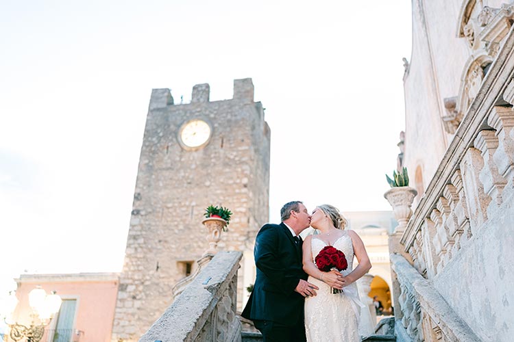 Bride and groom kissing on a stone staircase in Taormina, with the medieval clock tower behind them during their romantic wedding in Sicily