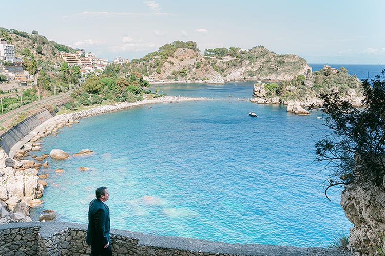 Groom standing on a stone terrace with panoramic views of Isola Bella and the Mediterranean during his romantic wedding in Taormina, Sicily.