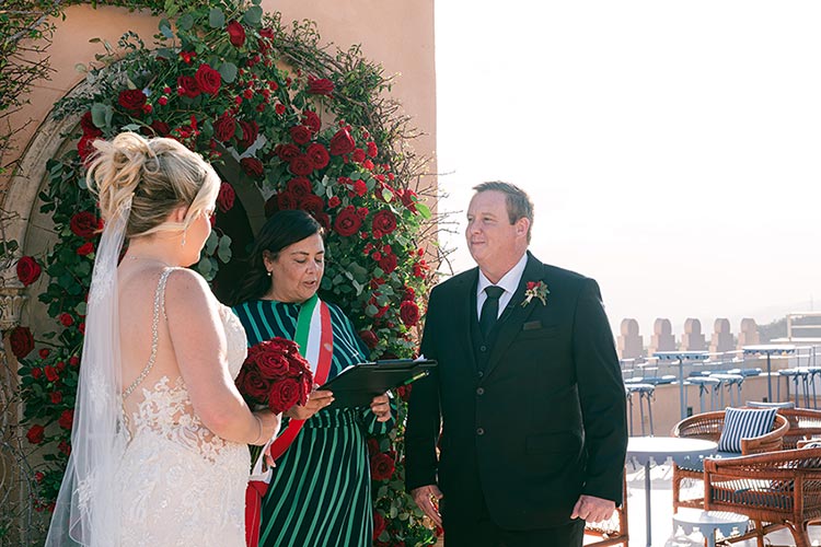 Wedding celebrant leading the symbolic ceremony in front of a red rose floral arch with the couple on a panoramic terrace in Taormina, Sicily.