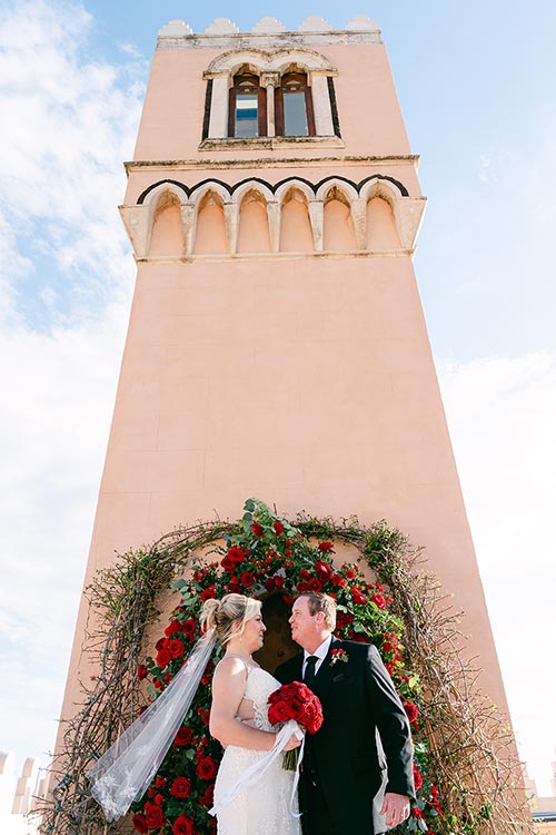 Bride and groom standing in front of a red rose wedding arch beneath a historic tower during their romantic ceremony in Taormina, Sicily.