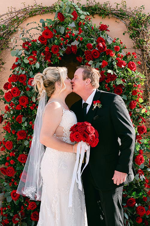 Bride and groom kissing in front of a red rose floral arch during their romantic wedding ceremony in Taormina, Sicily.