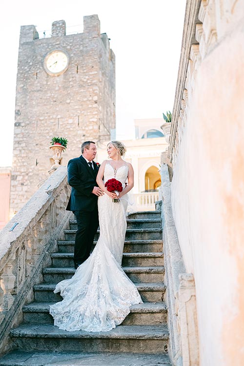 Bride and groom standing on a stone staircase with Taormina’s medieval clock tower behind them during their romantic wedding photoshoot in Sicily.