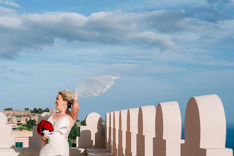 Bride holding her veil in the wind on a panoramic terrace overlooking the Mediterranean Sea during her romantic wedding in Taormina, Sicily.