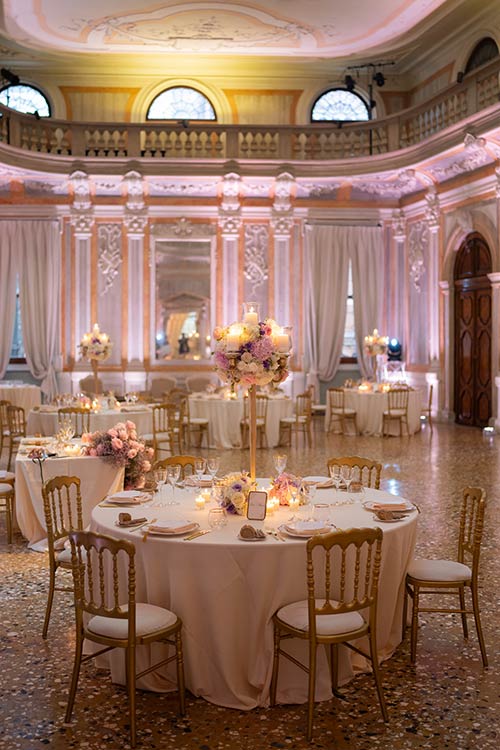 Elegant wedding dinner setup inside a historic Venetian palace with floral centerpieces and warm lighting.