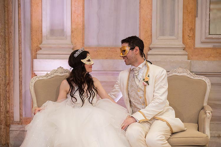 Bride and groom sitting together wearing Venetian masks and enjoying a quiet moment during their wedding reception.