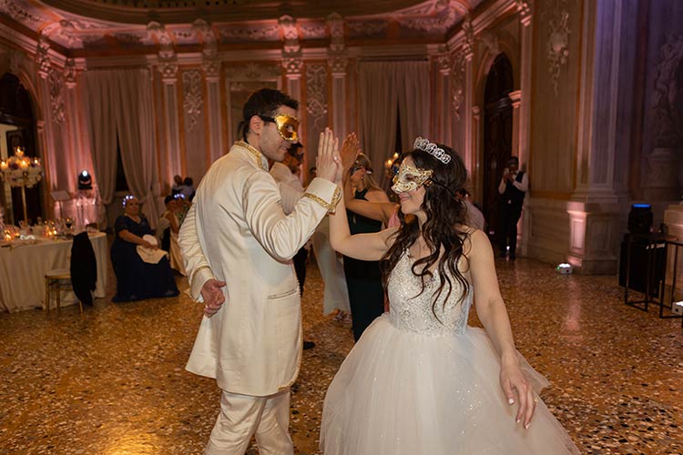Bride and groom dancing with Venetian masks during their wedding celebration in an elegant palace hall.