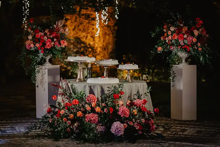 Romantic wedding cake display surrounded by colourful floral arrangements and evening lights